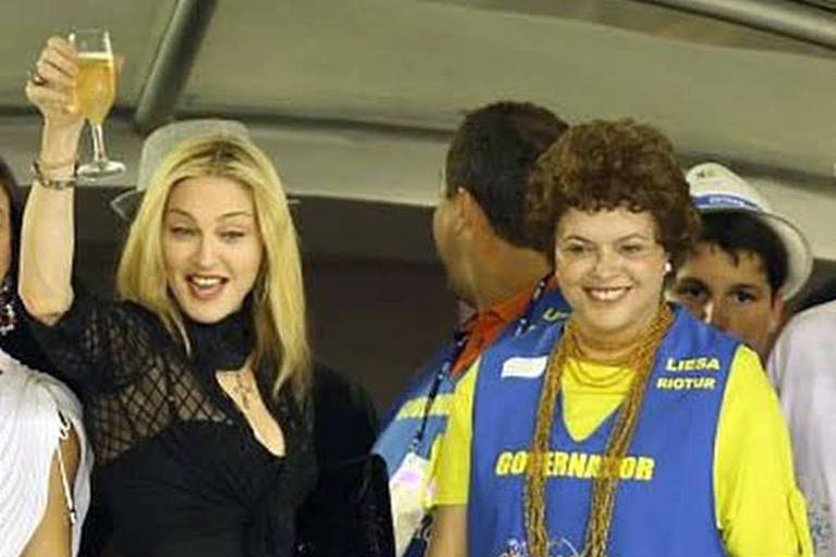A group of people is gathered at an event, with one woman raising a glass in a toast. She has long blonde hair and is wearing a black outfit. Next to her, another woman is smiling and wearing a blue vest with yellow accents, indicating a role related to the event. The background shows other attendees and a festive atmosphere.