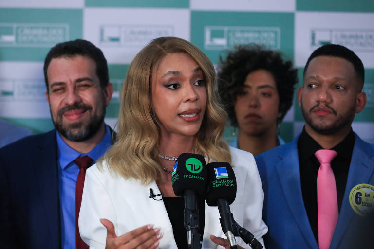 Uma mulher com cabelo loiro e liso， vestindo um blazer branco， fala ao microfone em uma coletiva de imprensa. Ao fundo， há três homens: um à esquerda com barba e cabelo curto， sorrindo; um no centro com cabelo cacheado e um à direita， usando um terno azul claro. O fundo é composto por painéis verdes com logotipos.