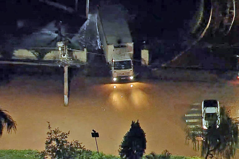 A imagem mostra uma cena noturna de uma área urbana inundada. Um caminhão está parcialmente submerso em água， enquanto outros veículos estão visíveis em uma estrada alagada. A iluminação dos faróis do caminhão reflete na superfície da água， que é de cor marrom. Ao fundo， há postes de luz e vegetação ao redor.