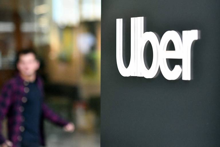 A imagem mostra o logotipo da Uber em destaque， com a palavra 039;Uber039; em letras brancas sobre um fundo escuro. Ao fundo， há uma pessoa desfocada， aparentemente saindo de um local.
