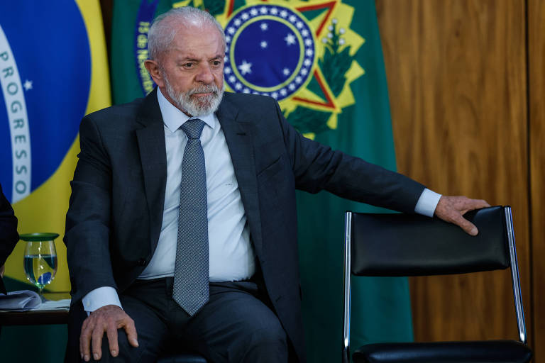 Um homem de cabelos grisalhos e barba， vestido com um terno escuro e gravata， está sentado em uma cadeira. Ele parece pensativo， com a mão apoiada na cadeira ao seu lado. Ao fundo， há bandeiras do Brasil e um símbolo nacional.