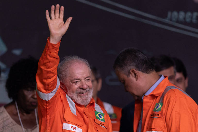 Um homem de cabelo grisalho e barba， vestindo um macacão laranja com detalhes brancos e a bandeira do Brasil， está acenando com a mão direita. Ele parece estar em um evento， cercado por outras pessoas que também estão vestindo macacões laranjas. O ambiente é de celebração.