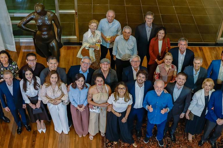 Uma grande reunião de pessoas em um ambiente interno， com piso de madeira e um tapete decorativo. Ao fundo， há esculturas em exibição. As pessoas estão dispostas em várias fileiras， algumas sorrindo e outras em poses descontraídas. A maioria está vestida de forma formal ou semi-formal， com uma variedade de cores e estilos. A iluminação é suave e há janelas grandes ao fundo.