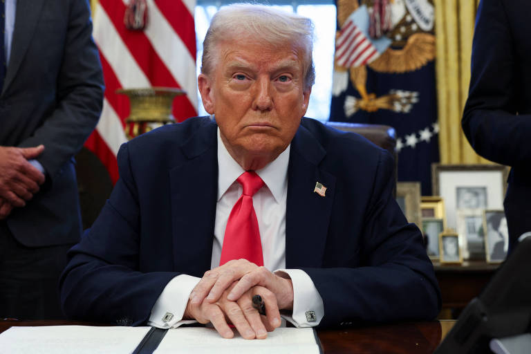 Um homem sentado à mesa em um escritório, com as mãos sobre um documento. Ele usa um terno escuro e uma gravata vermelha. Ao fundo, há bandeiras e outros objetos decorativos típicos do Escritório Oval.