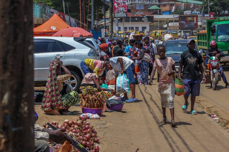 A imagem mostra uma movimentada rua de mercado， onde várias pessoas estão vendendo e comprando produtos. À esquerda， há uma pessoa vendendo cebolas e outros vegetais. No centro， algumas pessoas estão agachadas， organizando mercadorias em cestos. À direita， um grupo de pessoas caminha， algumas carregando sacolas. Ao fundo， há carros estacionados e uma atmosfera urbana com várias barracas e lojas visíveis.