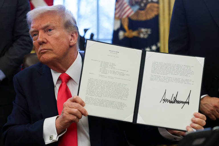 A imagem mostra Trump， um homem branco com cabelo loiro e uma gravata vermelha， sentado em uma mesa. Ele está segurando um documento em uma pasta， exibindo sua assinatura. Ao fundo， há uma janela com cortinas e bandeiras dos Estados Unidos.