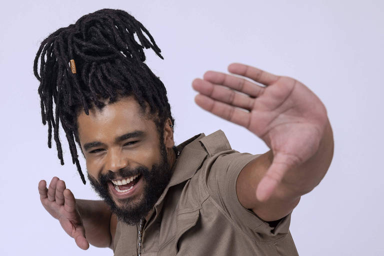 Um homem com cabelo dreadlock e barba está sorrindo e fazendo uma pose dinâmica, com os braços levantados e uma expressão alegre. Ele veste um macacão de cor marrom claro e está posicionado em um fundo branco.