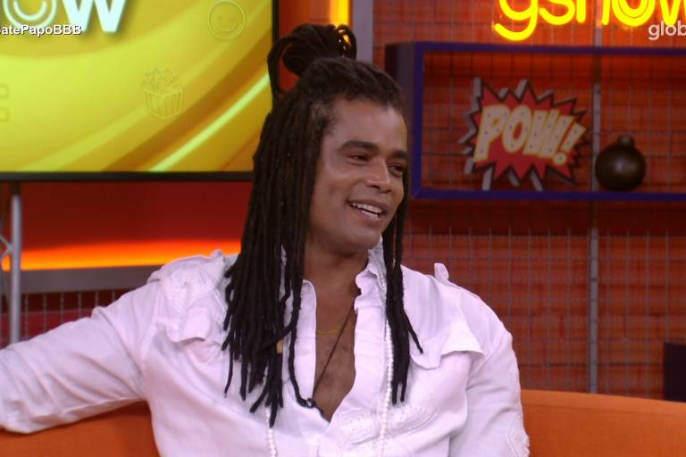 Um homem com cabelo longo e dreadlocks está sentado em um sofá laranja, sorrindo. Ele usa uma camisa branca com um decote em V. Ao fundo, há uma tela com o logo do programa e elementos decorativos coloridos, incluindo uma figura com a palavra 'POW'.