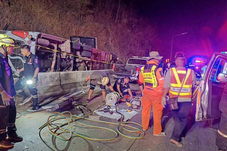 A imagem mostra um acidente de trânsito à noite， com um ônibus tombado ao lado da estrada. Várias pessoas， incluindo equipes de resgate e policiais， estão trabalhando na cena. Há equipamentos de resgate， como mangueiras e uma serra， espalhados pelo chão. Veículos de emergência estão presentes， com luzes piscando， e a vegetação é visível ao fundo.