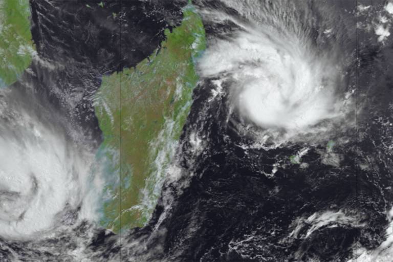 A imagem mostra uma vista do Oceano Índico, com duas tempestades visíveis à esquerda e à direita. À esquerda, uma tempestade está localizada próxima à costa de ilha, enquanto à direita, outra tempestade é visível em alto-mar. A parte superior da imagem exibe nuvens escuras e turbulentas, enquanto a terra firme aparece em verde.