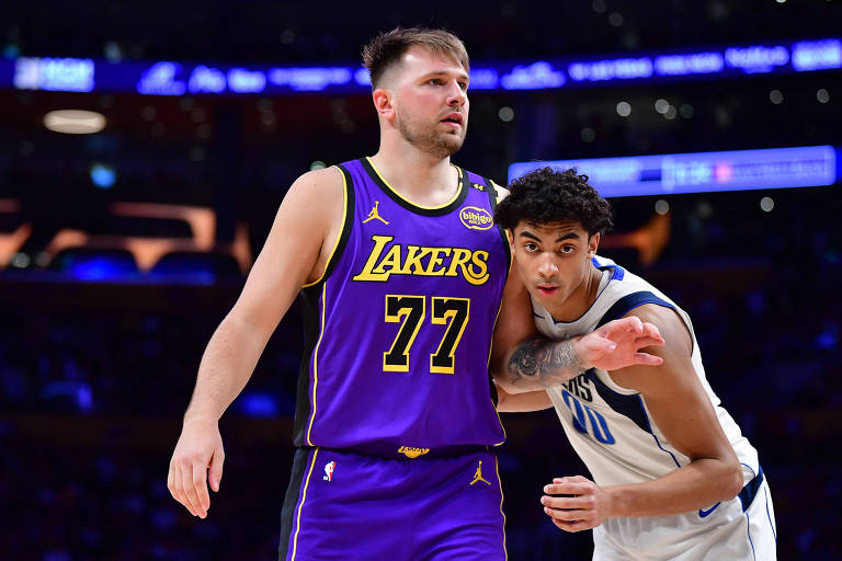 Dois jogadores de basquete estão em uma partida. Um deles está vestindo um uniforme roxo e amarelo dos Lakers， com o número 77， e parece estar em movimento. O outro jogador está usando um uniforme branco e azul， com o número 0， e está próximo ao primeiro jogador， tentando defendê-lo. O fundo mostra uma quadra de basquete iluminada.