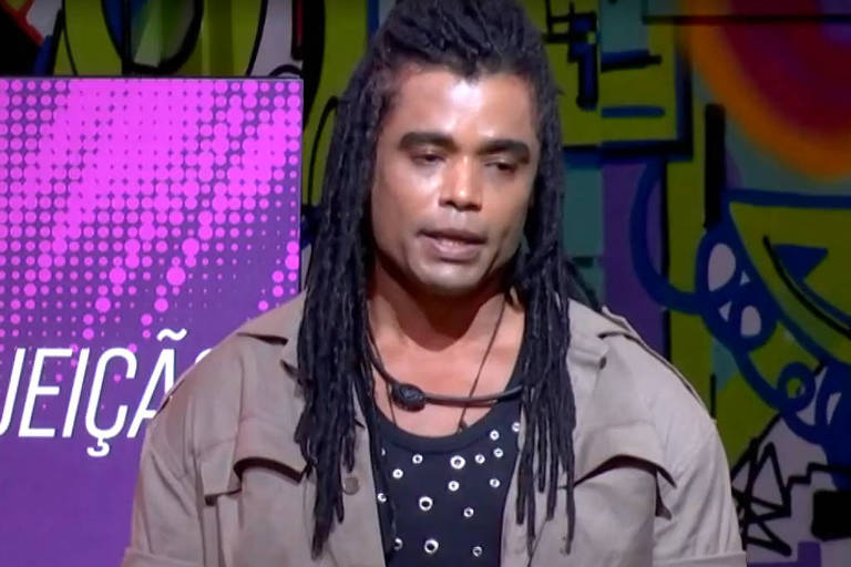 Um homem de cabelo longo e dreadlocks está em pé, com uma expressão séria, em frente a um grande painel que exibe a frase 'ELIMINADO(A) COM REJEIÇÃO'. O fundo é colorido e vibrante, com elementos gráficos. Na parte inferior da tela, há um contador que mostra o número 31 e a indicação 'AO VIVO'.