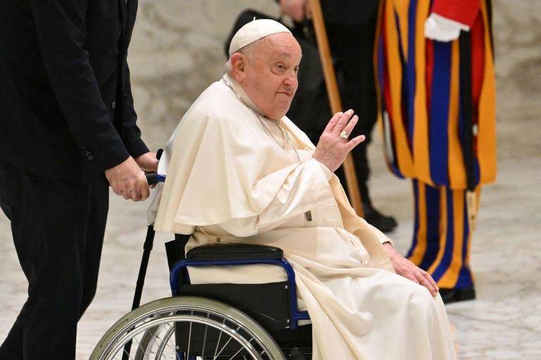 Imagem do Papa Francisco sentado em uma cadeira de rodas, acenando com a mão. Ele está vestido com uma túnica branca e uma estola. Um assistente está ao seu lado, empurrando a cadeira. Ao fundo, há membros da Guarda Suíça, vestidos com uniformes coloridos.