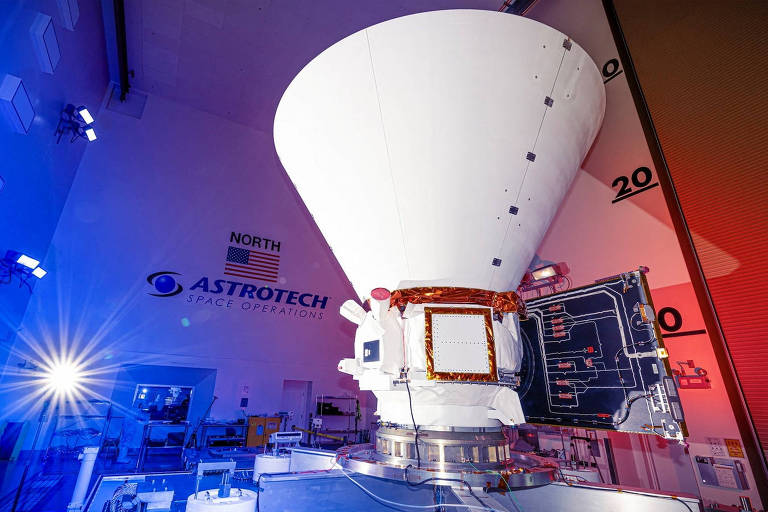 A imagem mostra uma nave espacial em uma instalação da Astrotech， com um grande cone branco na parte superior. O ambiente é iluminado com luzes azuis e vermelhas， e há uma parede com a marca 039;ASTROTECH SPACE OPERATIONS039;. No fundo， há uma escala numérica visível na parede.