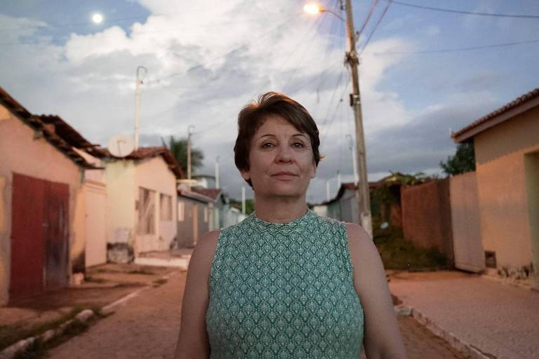 Uma mulher de cabelo curto e castanho， vestindo um vestido verde com padrões， está em pé em uma rua de um bairro. Ao fundo， há casas de cores claras e um céu nublado com a lua visível. A iluminação é suave， com postes de luz ao longo da rua.