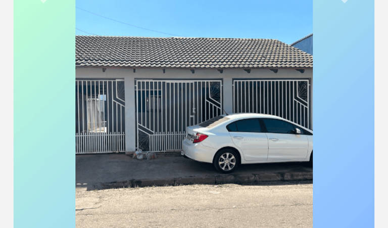 A imagem mostra a fachada de uma casa com um carro branco estacionado ao lado. A casa possui uma grade na frente, com janelas visíveis. O telhado é de telhas e o céu está claro, sem nuvens.