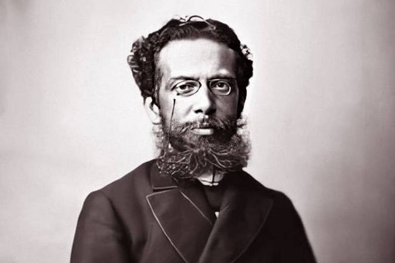 Fotografia em preto e branco é retrato de Machado de Assis , visto da cintura para cima. ele é um homem negro, de cabelos pretos curtos e ondulados. ele tem uma barba preta espessa. 