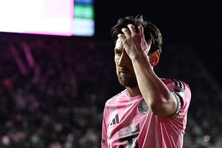 Um jogador de futebol está em campo， vestindo uma camisa rosa com detalhes em preto. Ele parece estar preocupado， passando a mão pela cabeça. O fundo é escuro， com algumas luzes e uma multidão visível.