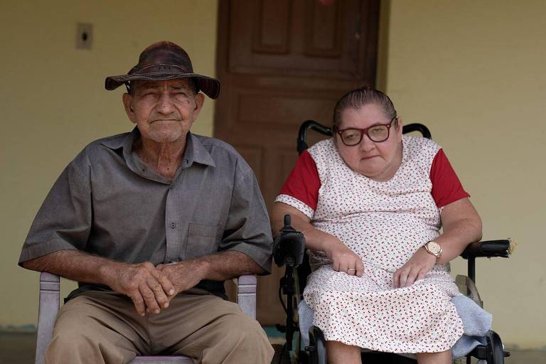 A imagem mostra um homem idoso e uma mulher idosa sentados em cadeiras. O homem usa um chapéu e uma camisa cinza， enquanto a mulher está em uma cadeira de rodas， vestindo um vestido branco com detalhes vermelhos e óculos. Ao fundo， há uma parede amarela e uma porta de madeira.