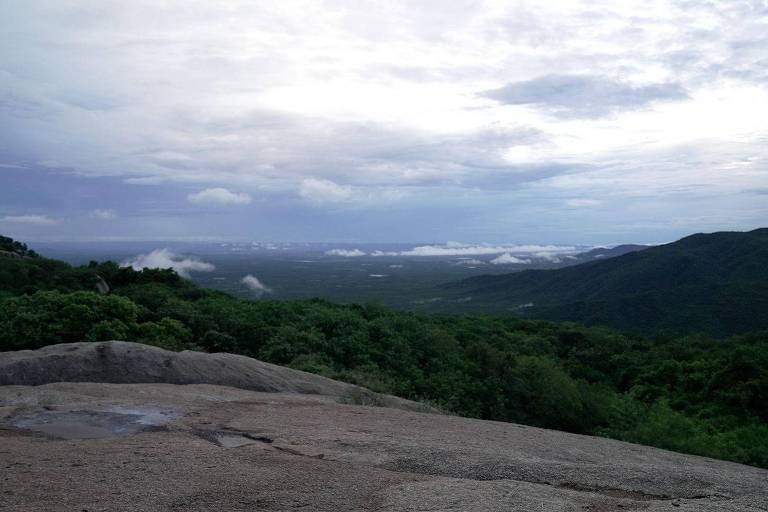 A imagem mostra uma vista panorâmica de uma paisagem montanhosa sob um céu nublado. No primeiro plano， há uma superfície rochosa e， ao fundo， uma vasta extensão de vegetação verde， com nuvens baixas cobrindo partes da paisagem.