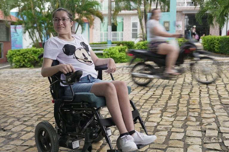 Uma jovem está sentada em uma cadeira de rodas motorizada em um calçamento de pedras. Ela usa uma camiseta branca com estampa do Mickey Mouse e shorts jeans. Ao fundo， há pessoas se movendo， incluindo uma pessoa em uma motocicleta. O ambiente é ao ar livre， com vegetação ao redor.