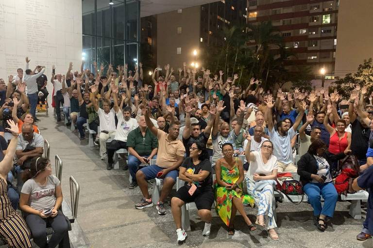 A imagem mostra uma grande reunião ao ar livre， onde um público numeroso está sentado em bancos. Muitas pessoas estão levantando as mãos， indicando participação ou votação. O ambiente é iluminado por luzes artificiais， e ao fundo， há prédios e árvores. A maioria das pessoas está vestida de maneira casual.