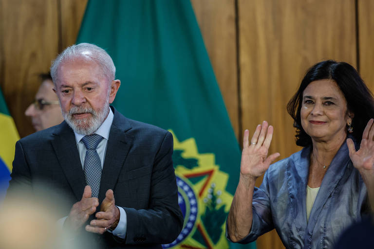 A imagem mostra duas pessoas em um evento oficial. À esquerda， um homem de cabelo grisalho e barba， vestindo um terno escuro e gravata， aplaude. À direita， uma mulher com cabelo escuro e solto， usando uma blusa clara e um casaco azul， acena com as duas mãos. Ao fundo， há bandeiras do Brasil e uma parede de madeira.