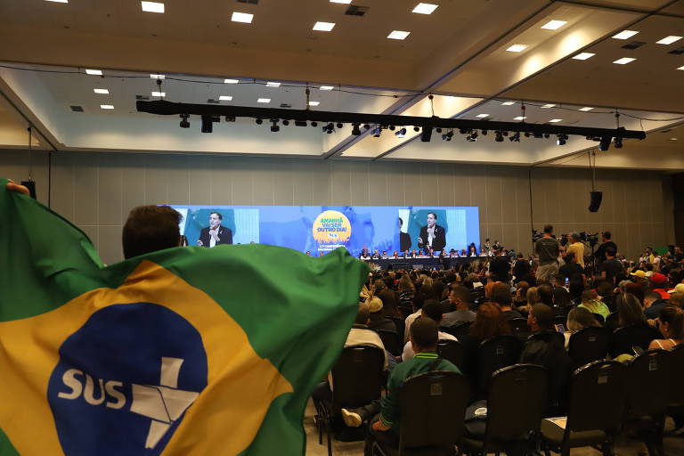 A imagem mostra uma grande sala de conferências com um público numeroso. No fundo， há um palco com uma mesa de autoridades e um telão exibindo um logo. Um homem na frente segura uma bandeira do Brasil com o símbolo do SUS. O ambiente é bem iluminado e as cadeiras estão ocupadas por pessoas que assistem ao evento.