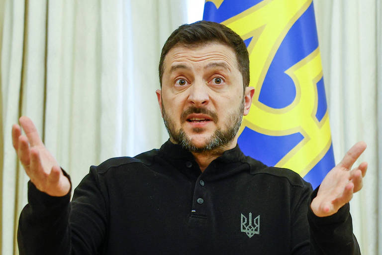 Um homem com cabelo curto e barba está em um discurso， gesticulando com as mãos. Ele usa uma camiseta preta com um símbolo que representa a Ucrânia. Ao fundo， há uma bandeira da Ucrânia com as cores azul e amarelo.