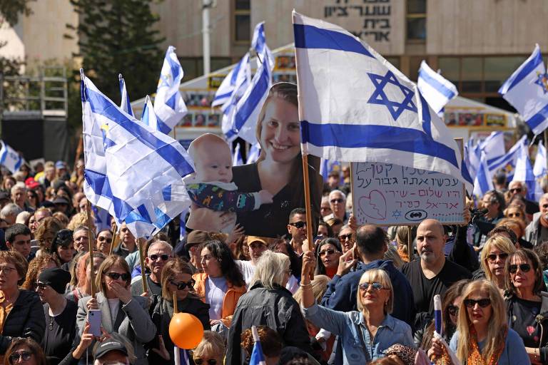 A imagem mostra uma grande multidão em uma manifestação， segurando bandeiras de Israel. Algumas pessoas estão segurando cartazes， e uma delas exibe uma foto de uma mulher segurando um bebê. O ambiente parece ser ao ar livre， com árvores ao fundo e um edifício visível.