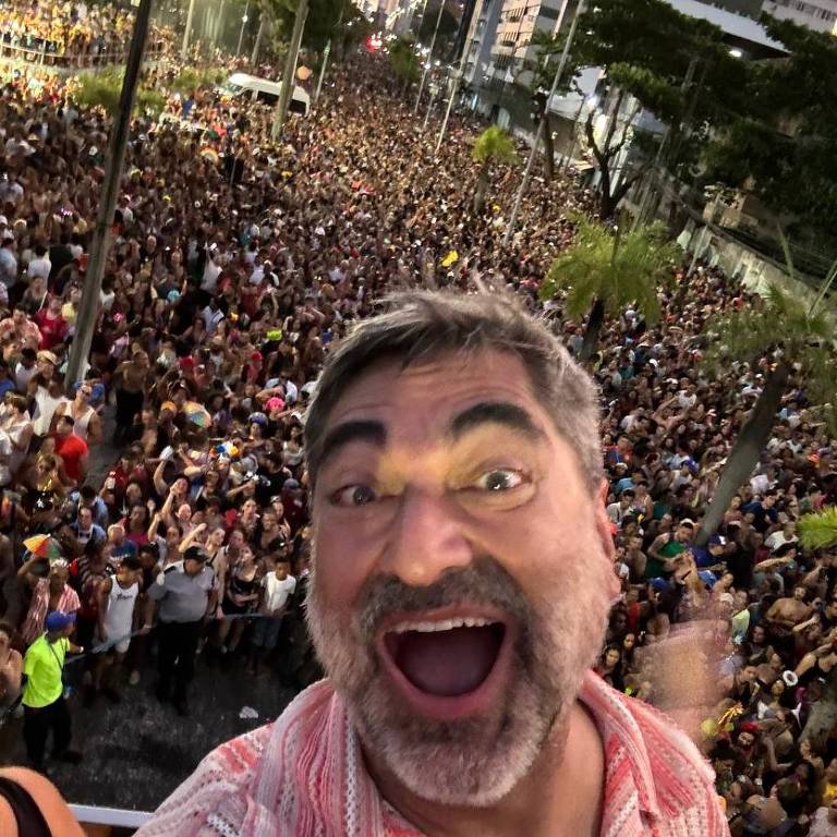 A imagem mostra um homem sorridente tirando uma selfie em um evento ao ar livre. Ao fundo， há uma grande multidão de pessoas reunidas， com árvores e prédios visíveis. O homem está usando uma camisa listrada e parece estar se divertindo.