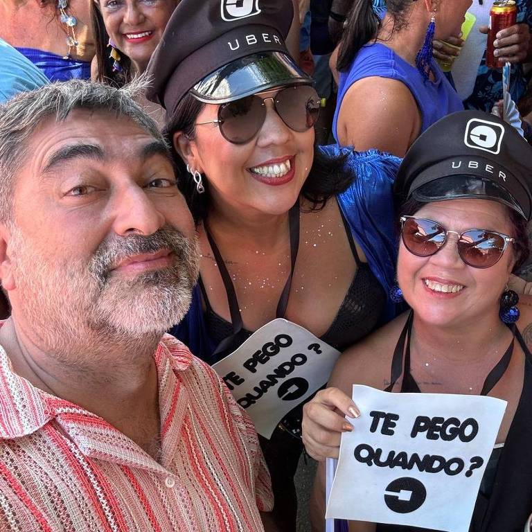 A imagem mostra um grupo de pessoas durante uma festa de Carnaval. Um homem está à frente， sorrindo， com uma camisa listrada. Ao seu lado， duas mulheres usam chapéus pretos com o logotipo da Uber e seguram placas brancas com a frase 039;TE PEGO QUANDO?039;. Ao fundo， há várias outras pessoas， todas vestidas de forma colorida e festiva.