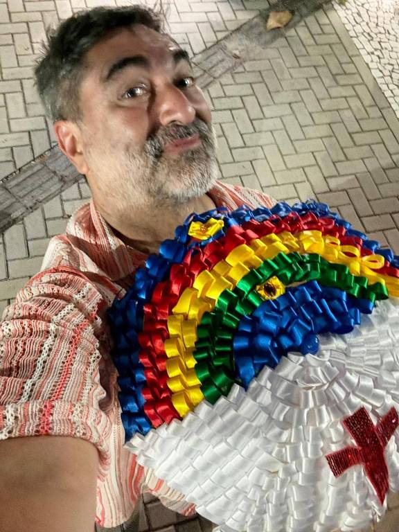 Um homem sorridente está tirando uma selfie. Ele usa uma camisa listrada e segura um grande adereço colorido em forma de arco-íris， que possui várias cores， incluindo azul， verde， amarelo e vermelho， além de um símbolo em vermelho. O fundo é uma calçada de pedras.