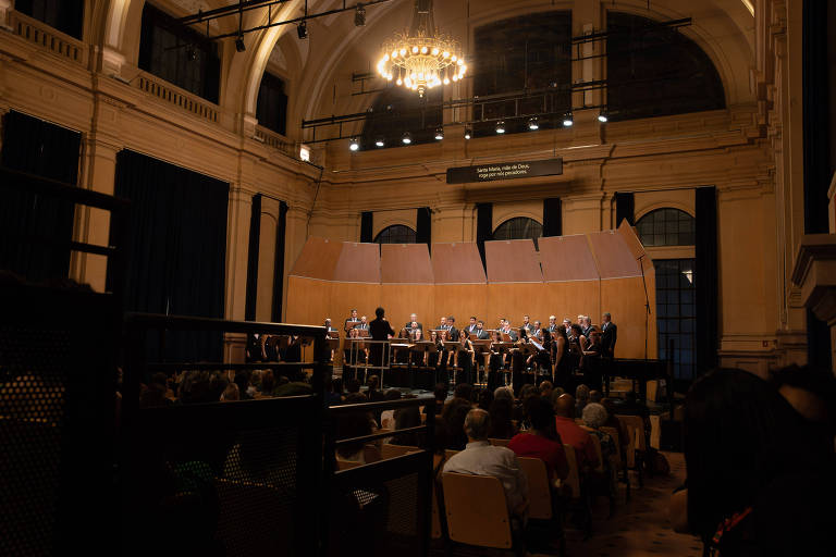 A imagem mostra um auditório com um grupo de músicos se apresentando em um palco elevado. O palco é cercado por painéis de madeira e há uma grande candelabro iluminando o espaço. A plateia， composta por várias pessoas， está sentada em cadeiras， assistindo à performance. O ambiente é bem iluminado， com janelas altas ao fundo.