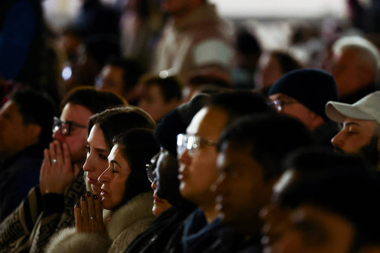 A imagem mostra um grupo de pessoas sentadas em um evento， com expressões de concentração e reflexão. Algumas pessoas estão com as mãos unidas em oração ou meditação， enquanto outras observam atentamente. O ambiente parece ser de uma reunião ou cerimônia， com iluminação suave e um fundo desfocado.