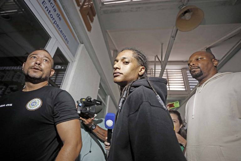 A imagem mostra três pessoas em um ambiente interno. À esquerda， um homem com uma camiseta preta e um emblema visível no peito， segurando uma câmera. No centro， uma mulher com cabelo trançado e usando um moletom preto， olhando para a câmera. À direita， um homem com uma camiseta clara， observando a cena. O ambiente parece ser um corredor com iluminação natural.