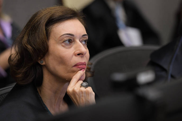 A imagem mostra uma mulher com cabelo castanho escuro e ondulado， usando um vestido preto. Ela está com a mão no queixo e parece estar prestando atenção em algo à sua frente