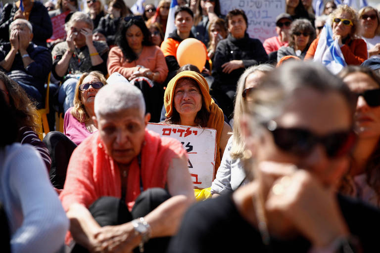 A imagem mostra um grupo de pessoas sentadas em um protesto. Em primeiro plano， há uma mulher com cabelo curto e grisalho， vestindo uma blusa vermelha e calças escuras. Ao fundo， uma mulher com um chapéu laranja e uma placa que contém texto em hebraico. O ambiente é de concentração， com várias pessoas ao redor， algumas com expressões sérias e outras olhando para frente.