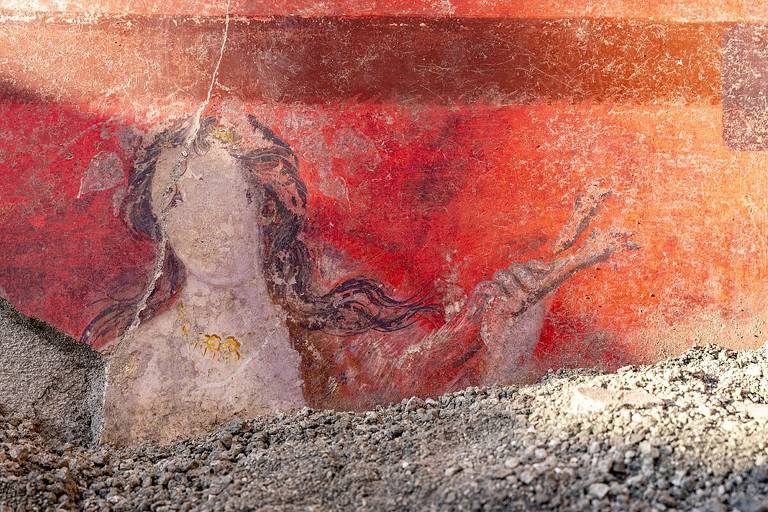 A imagem mostra um fragmento de um fresco antigo， com uma figura feminina parcialmente visível. A figura tem cabelos longos e ondulados e está segurando algo com a mão direita. O fundo é predominantemente vermelho， e a parte inferior da imagem está coberta por uma camada de cascalho e terra.