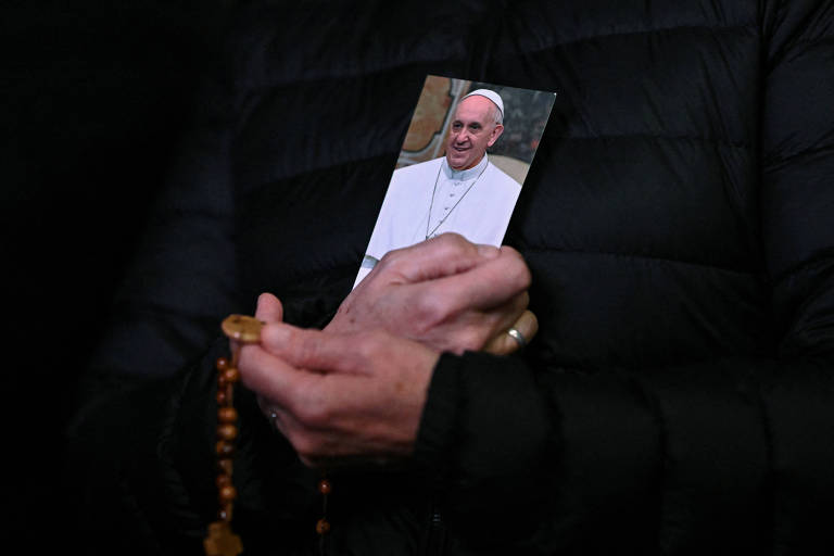 Uma pessoa está segurando uma foto do Papa Francisco， que está vestindo uma batina branca e sorrindo. A mão da pessoa também segura um terço， que é de contas marrons. A pessoa está vestindo uma jaqueta preta e o fundo da imagem é escuro.