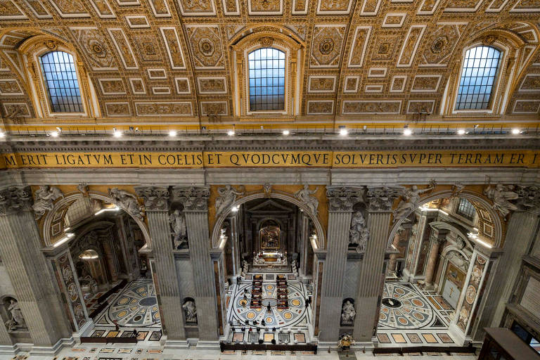 A imagem mostra uma vista panorâmica do interior da Basílica de São Pedro, em Roma. O teto é ricamente decorado com detalhes em dourado e há janelas altas que permitem a entrada de luz natural. As colunas são imponentes e há um piso com padrões geométricos. Na parte superior da imagem, há uma inscrição em latim que diz: 'MERIT LIGATVM ET IN COELIS ET QVODCVMQVE SOLVERIS SUPER TERRAM'.