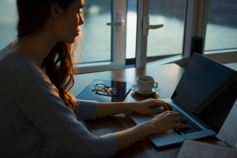 Uma mulher está sentada em uma mesa de madeira， digitando em um laptop. Ela tem cabelo longo e solto， e a luz do sol entra pela janela ao fundo， iluminando parcialmente seu rosto. Ao lado do laptop， há um copo de café e um par de óculos. A mesa também tem um tablet e alguns papéis.