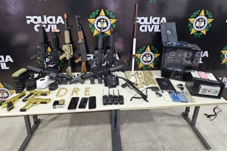 A imagem mostra uma mesa com diversos itens apreendidos pela Polícia Civil do Rio de Janeiro. Em cima da mesa， há várias armas falsas， incluindo rifles e pistolas， além de munições， celulares e outros objetos. Ao fundo， uma parede com o logotipo da Polícia Civil e a frase 039;Estado do Rio de Janeiro039;.