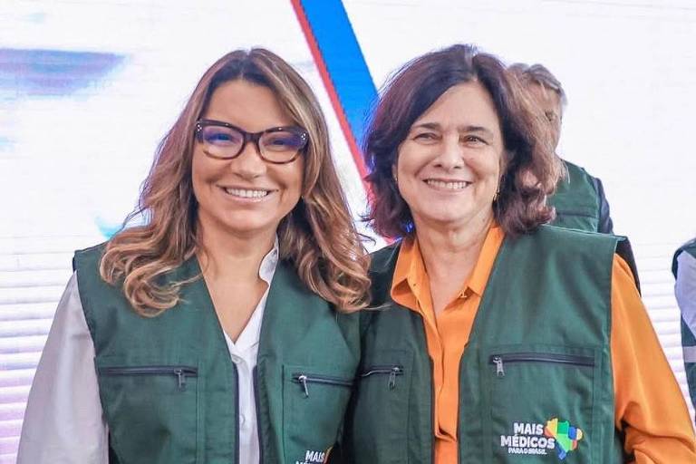 Duas mulheres sorrindo em um evento， vestindo coletes verdes com faixas refletivas. A mulher à esquerda usa óculos e uma blusa clara， enquanto a mulher à direita veste uma camisa laranja. Ao fundo， há um painel com o logo do SUS e do Ministério da Saúde.