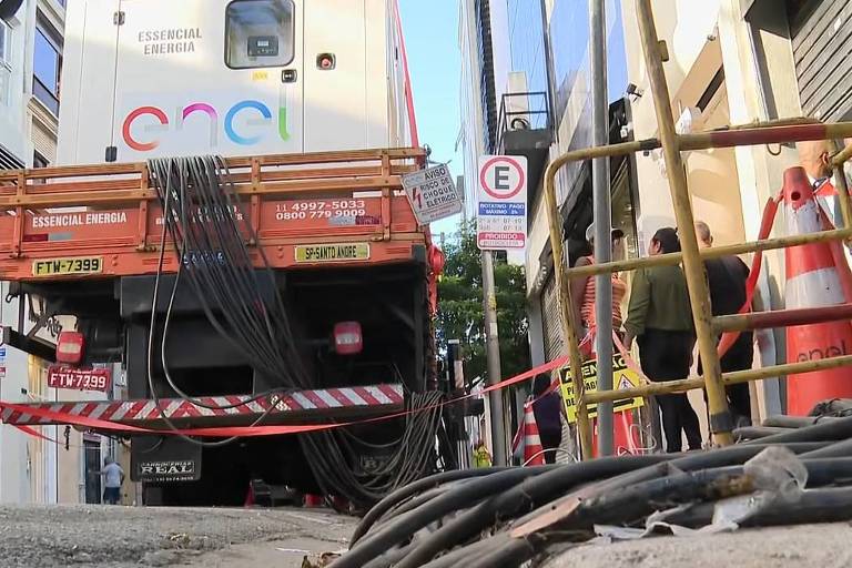 A imagem mostra a parte traseira de um caminhão de obras estacionado em uma rua urbana. O caminhão possui uma estrutura de madeira na parte de trás， com cabos elétricos se estendendo pelo chão. Há cones de sinalização e fita de advertência ao redor do caminhão. Ao fundo， é possível ver prédios e pessoas caminhando. Algumas placas de estacionamento estão visíveis.