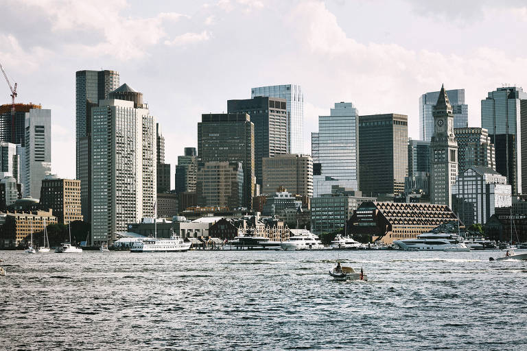 A imagem mostra uma vista panorâmica de uma cidade moderna com arranha-céus e edifícios altos. No primeiro plano， há um corpo d039;água com vários barcos e iates. O céu está parcialmente nublado， e a cidade apresenta uma mistura de arquitetura contemporânea e clássica， com destaque para um edifício com um relógio visível.