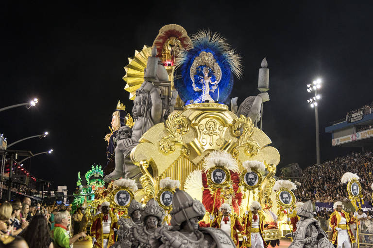 A imagem mostra um carro alegórico do Carnaval， ricamente decorado com elementos dourados e coloridos. No topo， há uma figura que parece ser uma deidade， cercada por penas e adornos. O carro é acompanhado por pessoas vestidas com trajes elaborados， e ao fundo， uma grande multidão observa o desfile sob as luzes do estádio.