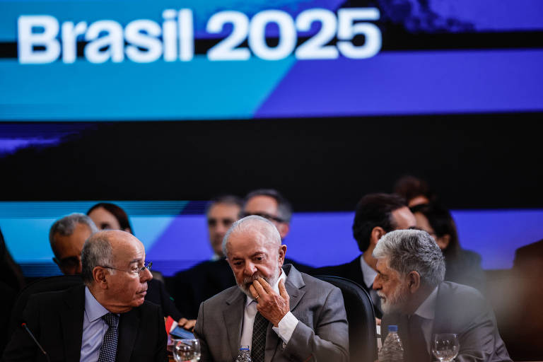 A imagem mostra uma reunião com três homens sentados à mesa， conversando entre si. Ao fundo， há um grande painel com a inscrição 039;Brasil 2025039;. Os homens estão vestidos formalmente， e há copos de vidro sobre a mesa. O ambiente parece ser de um evento oficial， com outras pessoas visíveis ao fundo.