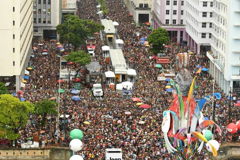 A imagem mostra uma grande multidão reunida em uma rua， com pessoas de diversas idades e estilos. Há barracas e tendas coloridas， além de veículos estacionados. Árvores e prédios cercam a área， e há uma decoração colorida na parte inferior da imagem， possivelmente relacionada ao evento. O clima parece festivo， com muitas pessoas interagindo.