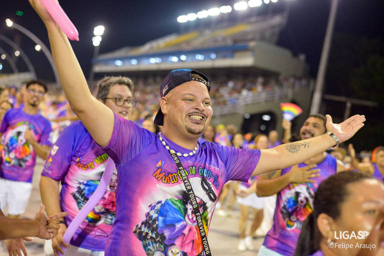 A imagem mostra um grupo de pessoas participando de um desfile de Carnaval. Um homem em destaque， com uma camiseta colorida e um boné， está sorrindo e levantando os braços em celebração. Ao fundo， há uma multidão e arquibancadas iluminadas， criando um ambiente festivo.